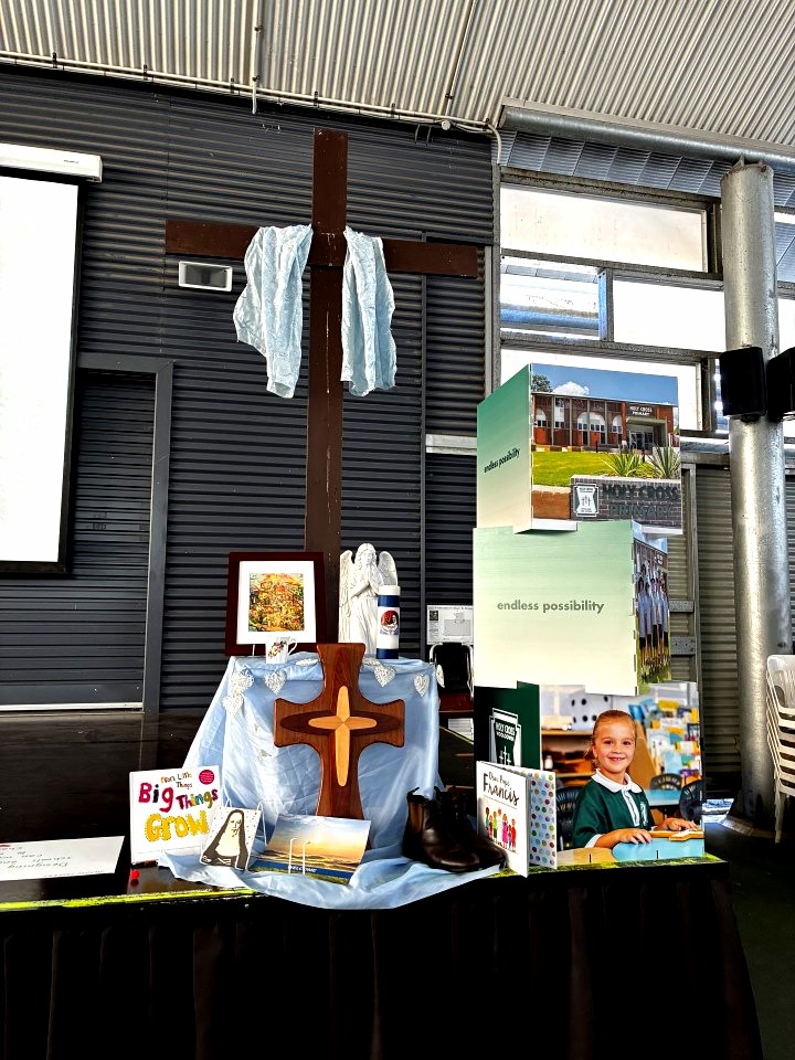 A Mercy Formation Day display featuring a large wooden cross draped with blue fabric, religious symbols, books and images representing Mercy values. The display is set up in a school hall and includes references to faith, community, learning and service.