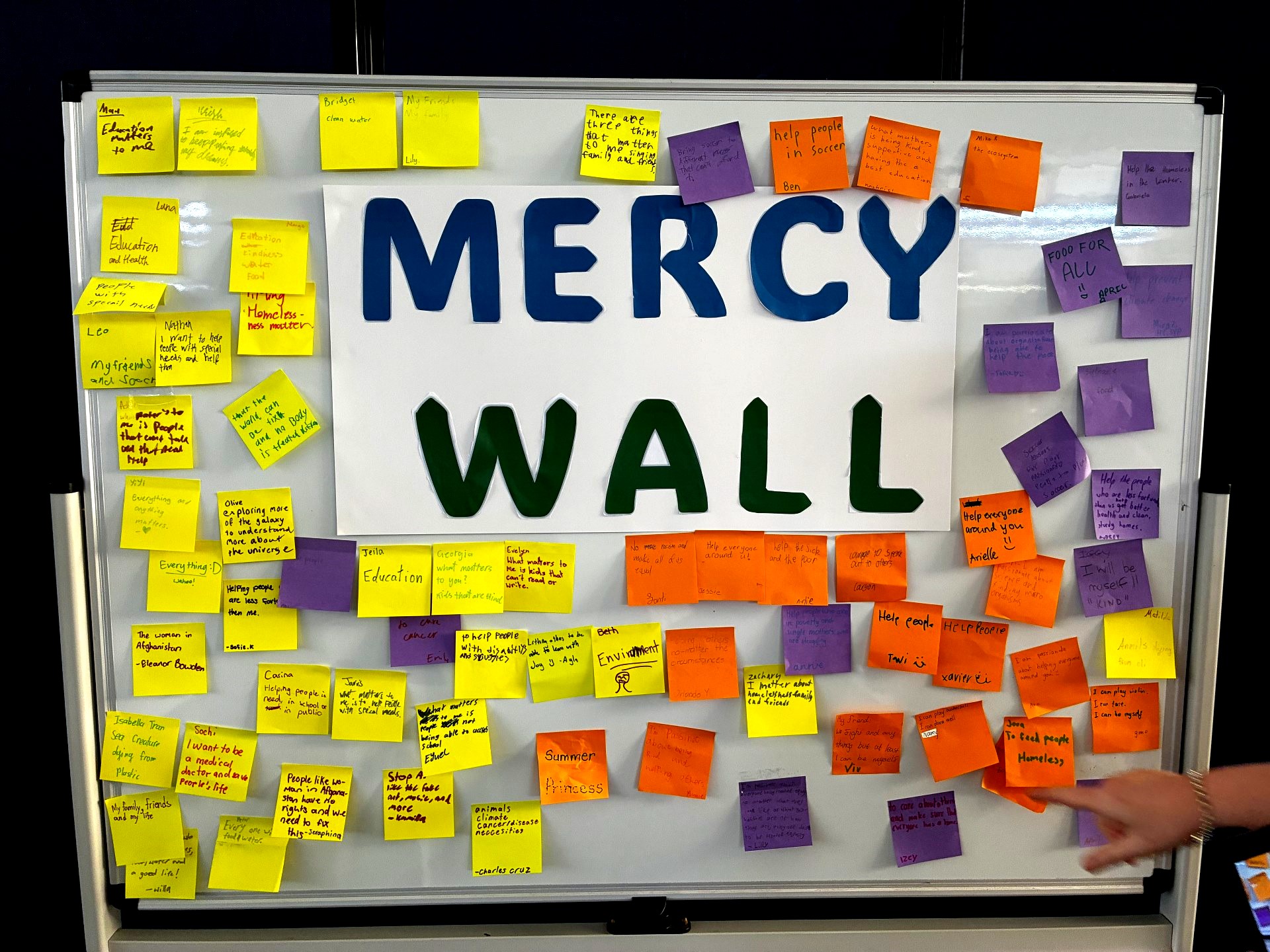 A display board titled "Mercy Wall" covered with colourful sticky notes written by students. The notes share ideas about kindness, helping others, caring for the environment and ways young people can make a positive impact in their communities.