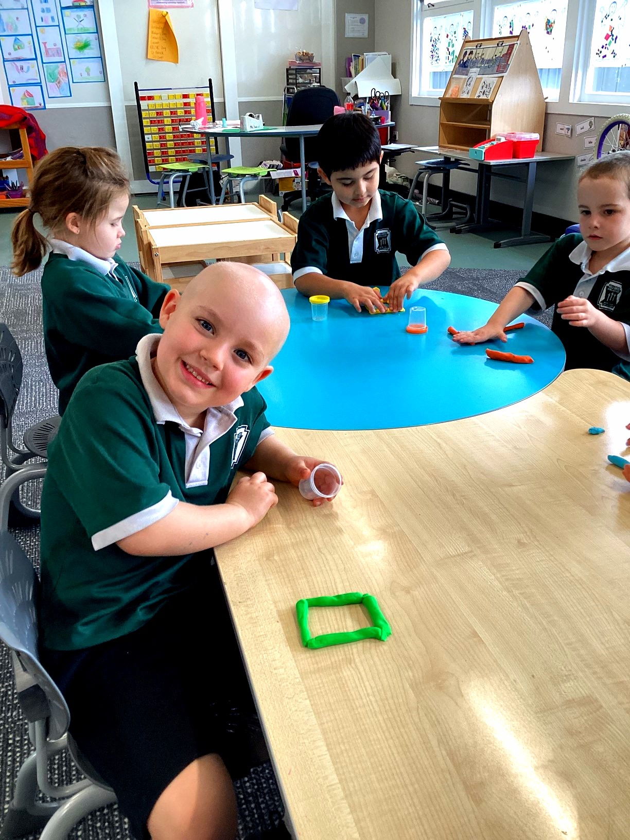 Prep students use modelling clay to create and explore 2D shapes during a hands-on mathematics lesson. A square shape made from green clay is displayed on the table in the foreground.