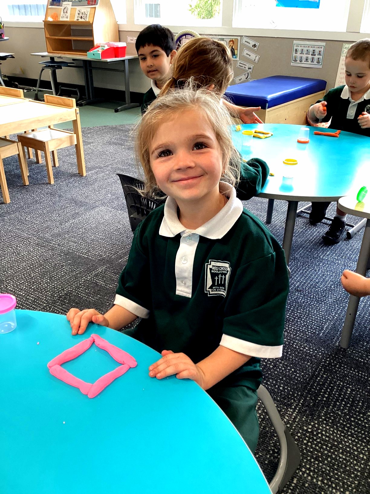 A Prep student sits beside a pink square shape made from modelling clay during a mathematics activity focused on recognising and describing 2D shapes.