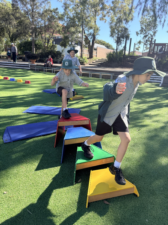 Students in school uniforms step and balance across a colourful outdoor obstacle course during a play-based activity.